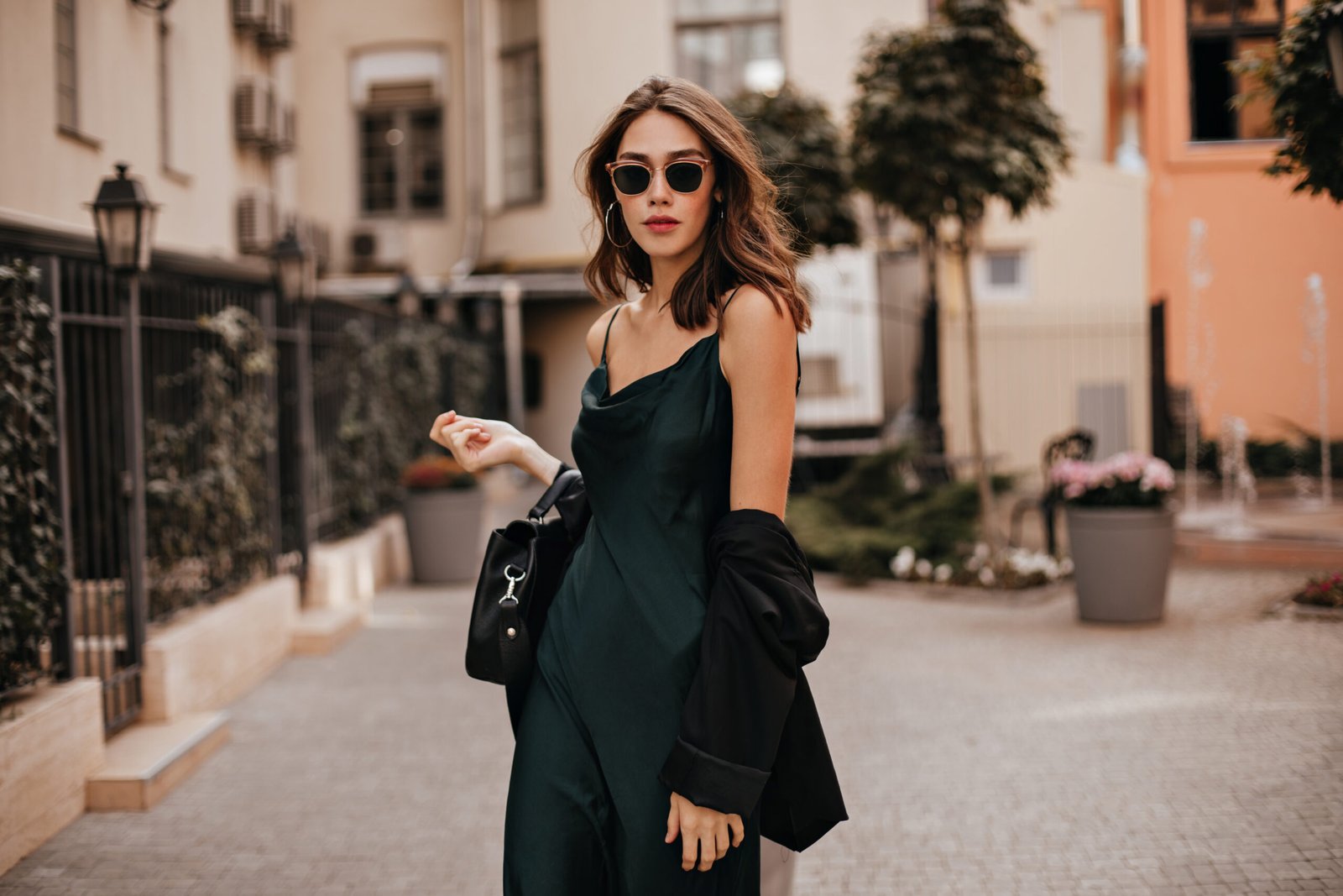 Fashionable pale brunette in long green dress, black jacket and sunglasses, standing on street duri Mode femme